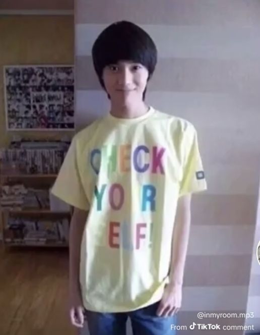 picture of taemin as a microbe wearing a pale yellow shirt with colorful letters that say "check yourself". he has a pleasant smile