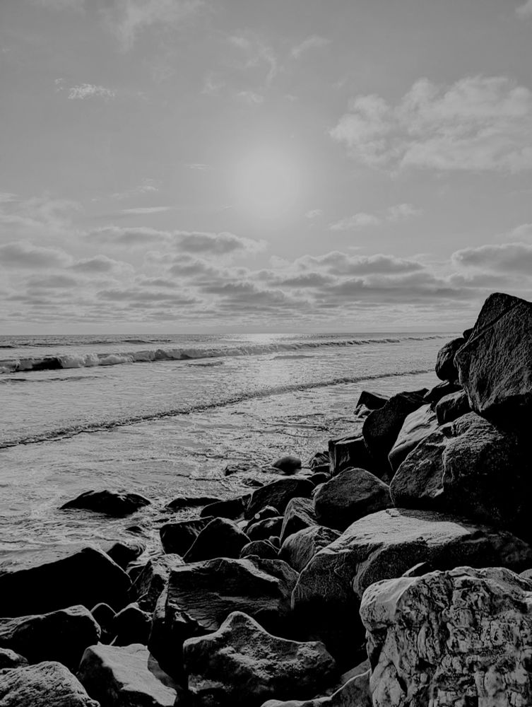 Greyscale photo of the sun over a rocky sea shore