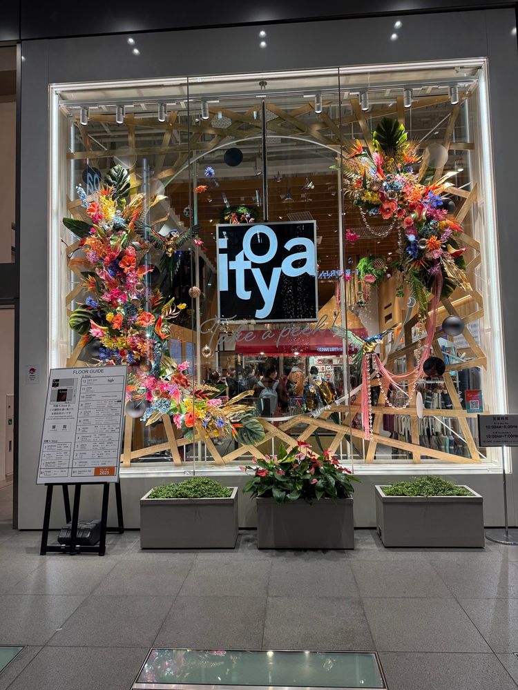 A picture of a large shop window of the Japanese store “Itoya”. The stylised logo of the store is surrounded by a large paper flowers mounted on a structure of wooden beams and columns. In front of the window is a small explanatory poster, a large square potted plant, and flanking it are two slightly shorter square pots of shrubs. 