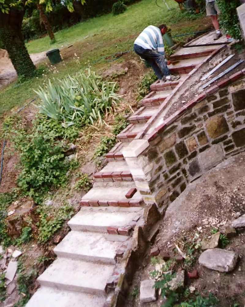 Des escaliers extérieurs en béton lavé avec un muret sur la droite. Un jardin sur la gauche. 