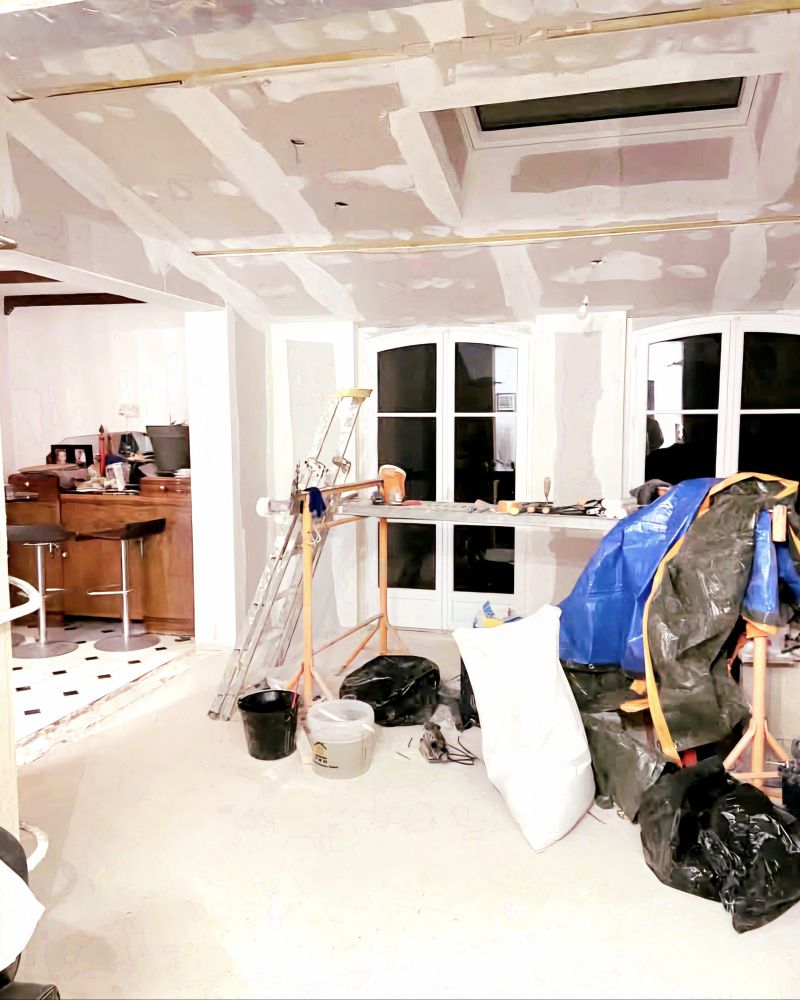 Une pièce en travaux avec de grandes fenêtres (il fait nuit dehors). Les murs sont en plaques de BA 13 avec les joints enduits. Au plafond il y a un grand velux. Dans la pièce il y a plein de bazar de chantier. Sur la gauche on aperçoit une pièce avec un meuble en bois et des tabourets de bar devant. 