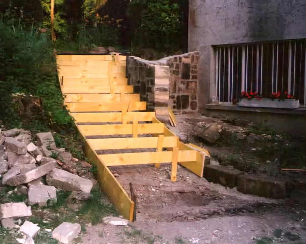 Des fondations en bois d'escaliers extérieurs avec un muret sur la droite et une maison. Un jardin au fond et sur la gauche avec un tas de blocs. 