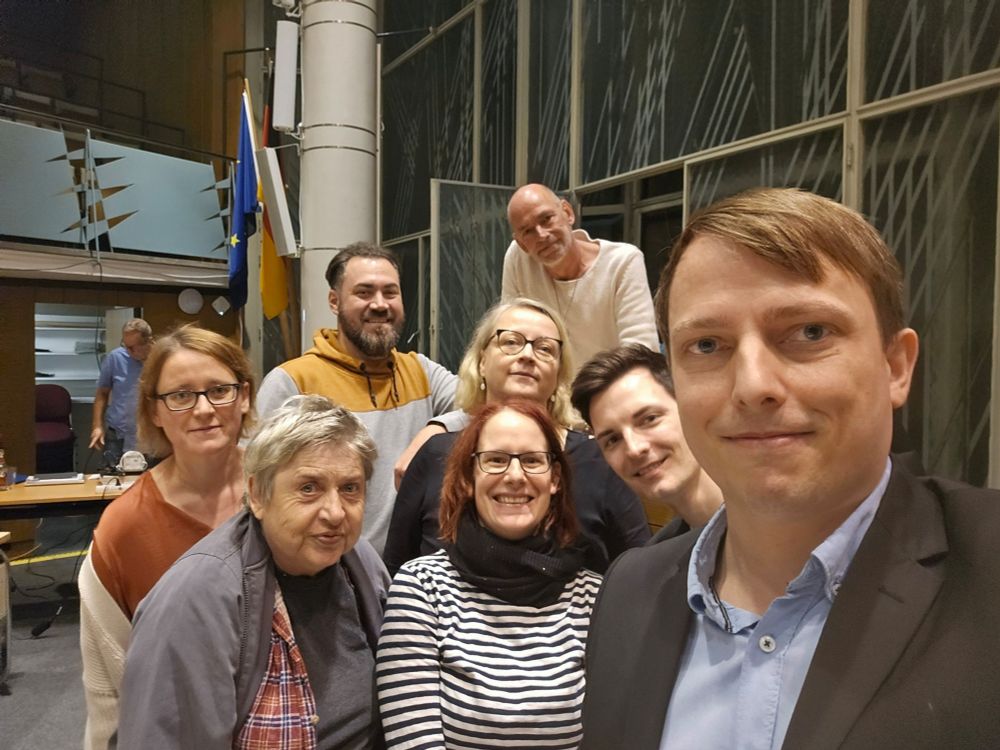 Ein Gruppenselfie von Teilen der Grünen Fraktion in der BVV Friedrichshain-Kreuzberg in einem Sitzungssaal mit Holztribüne und geschliffenen Glasfenstern.