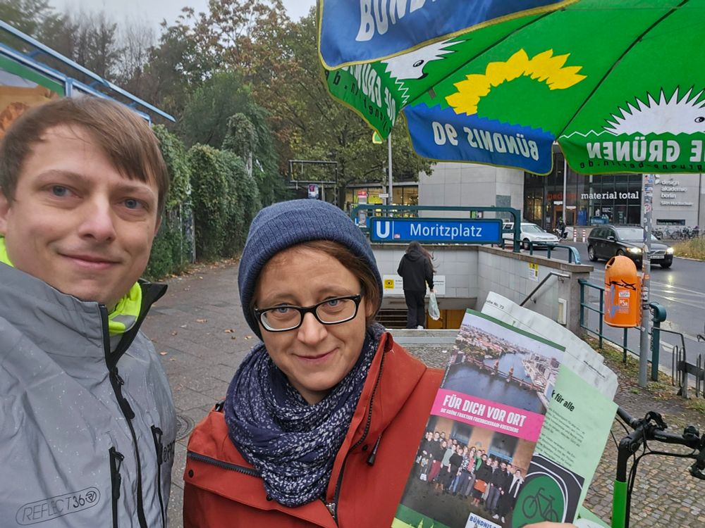 Ein Selfie mit Olja Koterewa und Pascal Striebel vor einem Lastenrad mit grünem Schirm. Im Hintergrund ein Eingang zur U-Bahn-Station Moritzplatz und ein Gemeinschaftsgarten mit grün bewachsenen Zäunen.