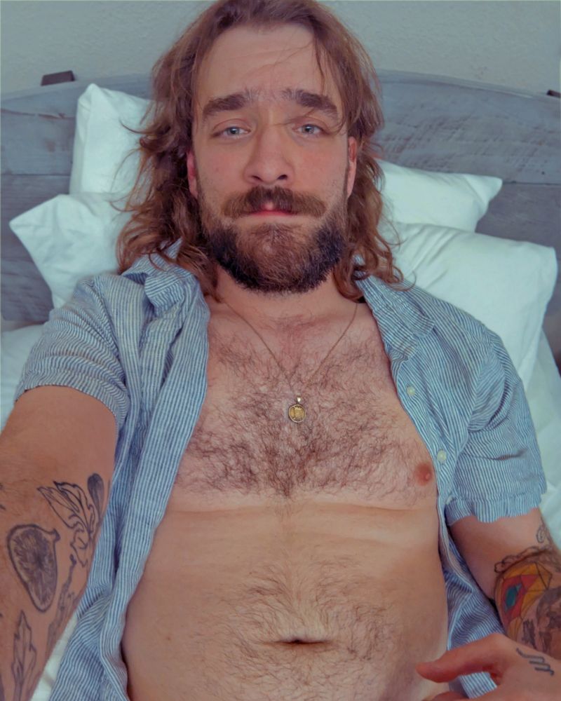 Photo of Hayden, a white man with shaggy brown hair and a scruffy beard, with tattoos on his forearms and hair on his chest and stomach. He is mostly undressed, wearing an unbuttoned striped linen shirt, lying back in bed with pillows and a headboard behind him. 