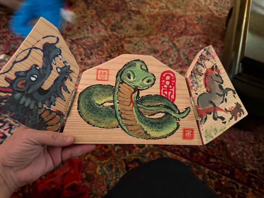 Hand-painted Buddhist ema (a votive plaque). When closed it depicts an orobourus dragon in green with wishes for health and happiness in the year (2024). 

When opened (see second photo) the plaque has three images, a triptych. On the left is another dragon (for the year 2024), in the middle is a friendly snake (for 2025), and on the right is a horse (2026, which will be the Year of the Horse.

Artist: Hank Tusinski. 