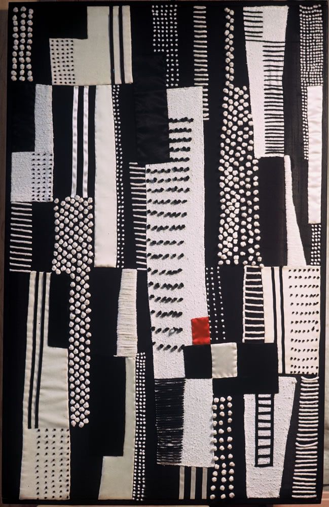 A 3 by 4 foot textile made of higglty jigglty blocks of back and white fabric, ribbon, and yarn french knots. A small, red, satiny rectangle 2/3 of the way down is its only color. 