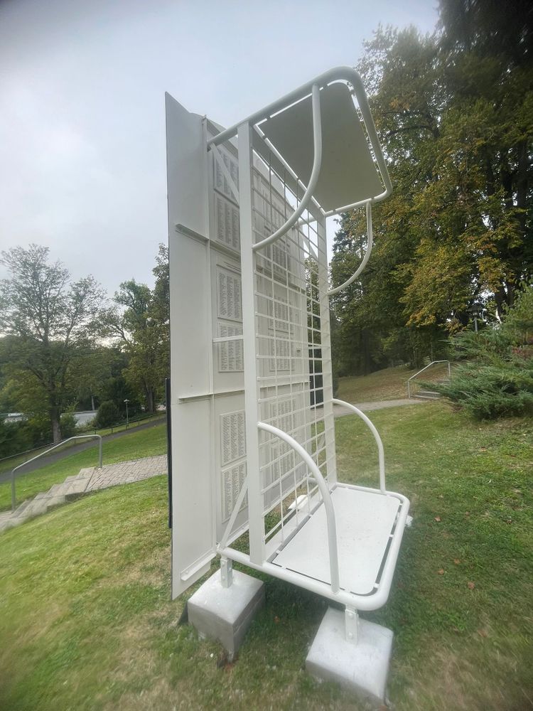 Denkmal der NS-'Euthanasie' in Weilmünster von vorne. Man sieht ein auf den Kopf gestelltes weißes Bett, das den vorderen Teil des Denkmals hält. Hinter dem Lattenrost sieht man in kleiner Schrift die Namen der im NS ermordeten Menschen.