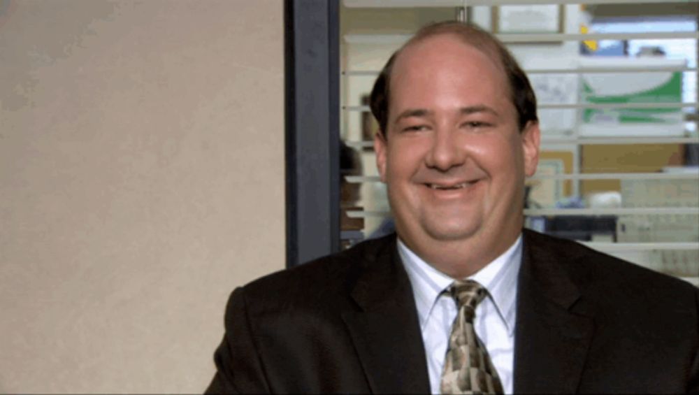 a bald man in a suit and tie smiles in front of a window