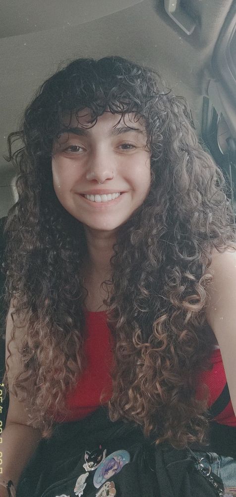 foto de uma mulher sorrindo de cabelos compridos e cacheados. ela usa uma regata vermelha e tem uma mochila preta sob o colo. 