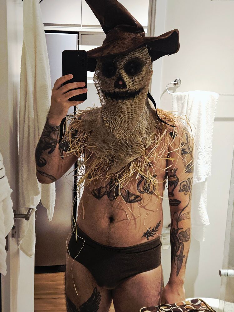 Selfie minha de cueca no espelho do banheiro. Estou de cueca e com uma fantasia de espantalho, uma colar de palha, uma máscara de juta com um rosto de espantalho preto e um chapeu pontudo marrom.