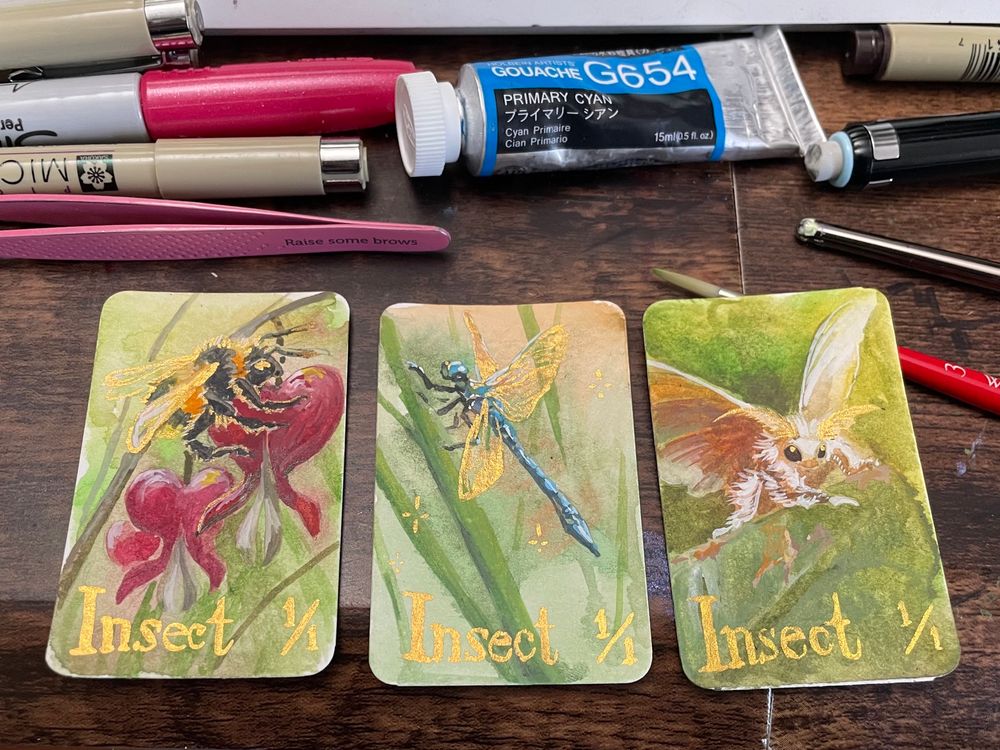 Three brightly colored miniature mtg tokens, each hand painted in watercolor and gouache, of 1/1 insects. The first is a bumblebee on a bleeding heart flower. The second is a blue dragonfly. The third is a fluffy white moth. All have some metallic gold accents.