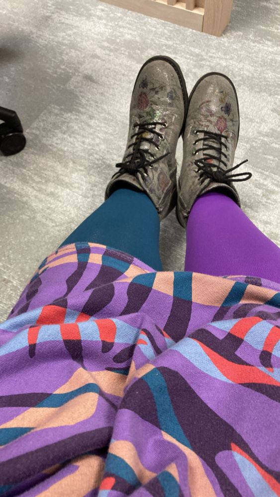 Picture of legs with multi-coloured dress - purple, green, red, blue and gold. Tights - left leg green, right lef purple. Grey boots and grey floor. 