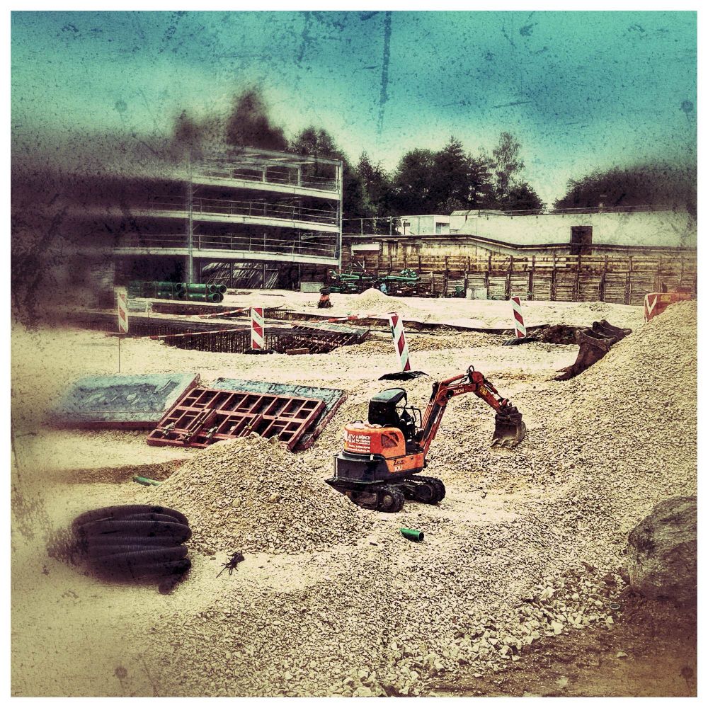 Minibagger auf einer Grossbaustelle zwischen zwei Kiesbergen, im Hintergrund der Rohbau eines Parkhauses.