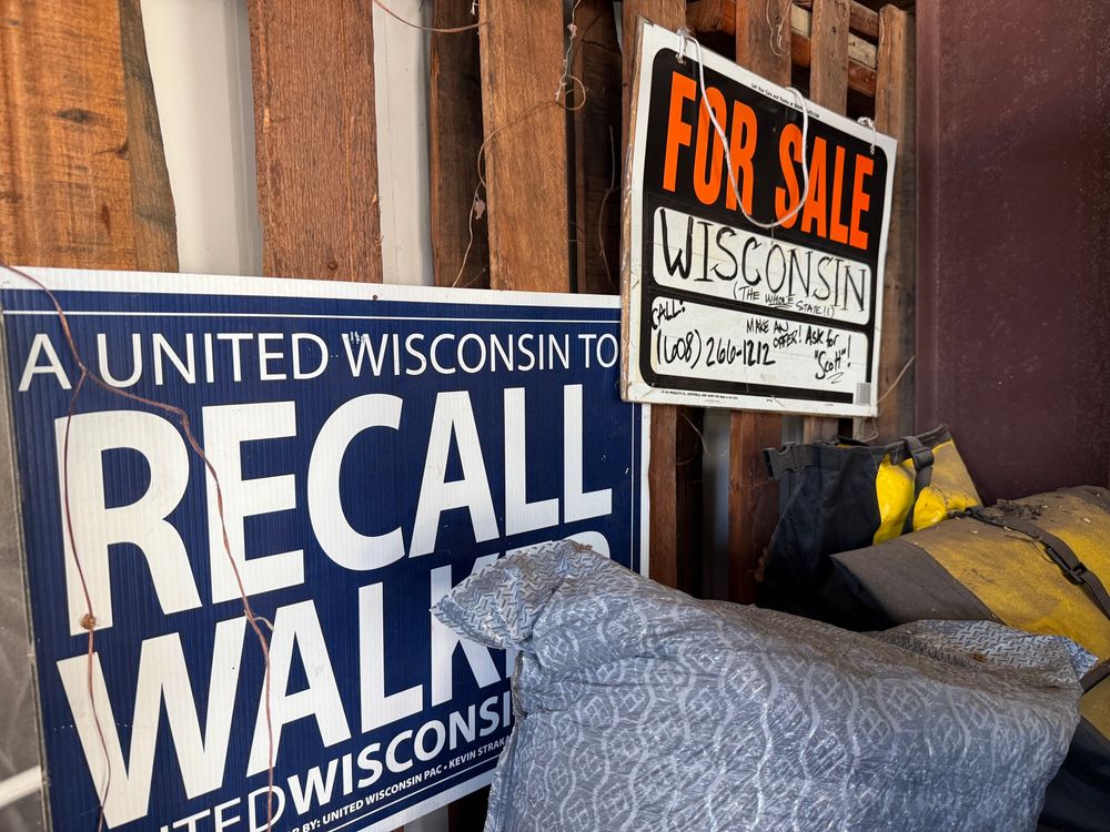 Two signs hang in a messy garage. One reads: “A united Wisconsin to RECALL WALKER.” A second reads: “FOR SALE: WISCONSIN.”