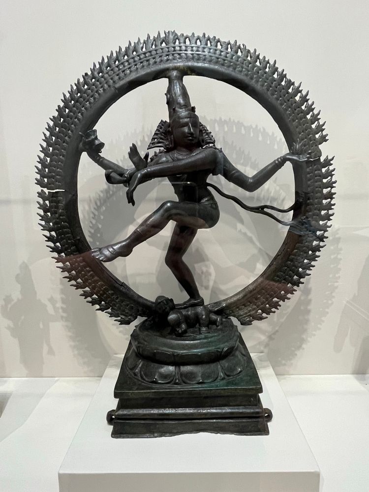 An iron statue of Nataraja (King of Dancers), one of many manifestations of the Hindu god Shiva. He is shown balancing on one leg as he lifts the other in his cosmic dance of creation and destruction of the universe.

In his upper right hand, he carries a drum representing creation; his upper left hand holds a flame symbolizing destruction. His two other hands are positioned in gestures of comfort. Surrounded by a flaming aureole, or circle of light, Shiva tramples on the demonic dwarf Apasmara, who personifies ignorance.

This sculpture is technically unusual because Shiva’s aureole was cast around a circular iron core. Such bronze images were carried around temples during festivals dedicated to Shiva. This object and the Chola bronzes on either side come from the region of Thanjavur, Tamil Nadu, in southeastern India.

Source: Saint Louis Art Museum 