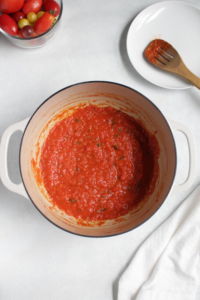 A small batch of tomato sauce in a pot. Next to the pot is a small bowl filled with ripening tomatoes and a white plate with a wooden spoon resting on it. The sauce was made with 1 pound of fresh tomatoes, a small onion, garlic clove, olive oil, basil, and a bay leaf.