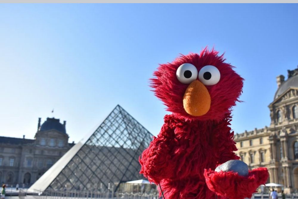 A drawing of Elmo from the sesame street franchise. He is a small red furry monster with a big orange nose is annoyed while holding a small grey rock named Rocco. The background is a blue sky with some buildings
