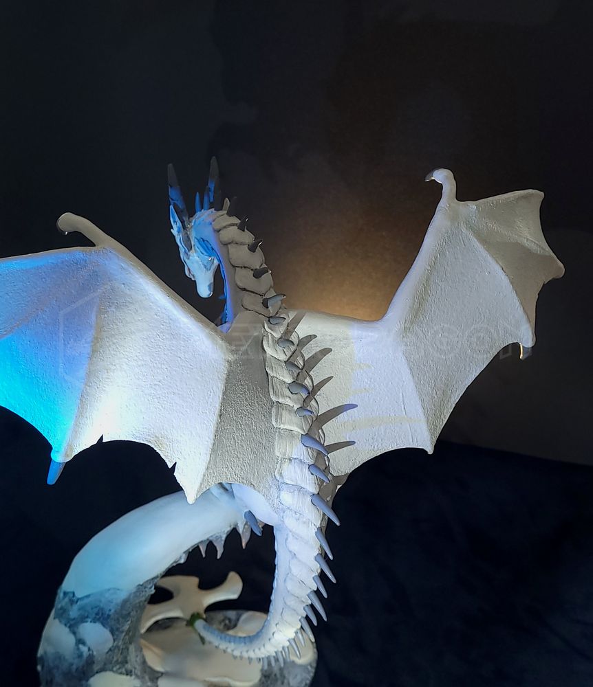 A modeled figurine of a white dragon sitting on a mountain ledge. Backside view with spines and wings visible.