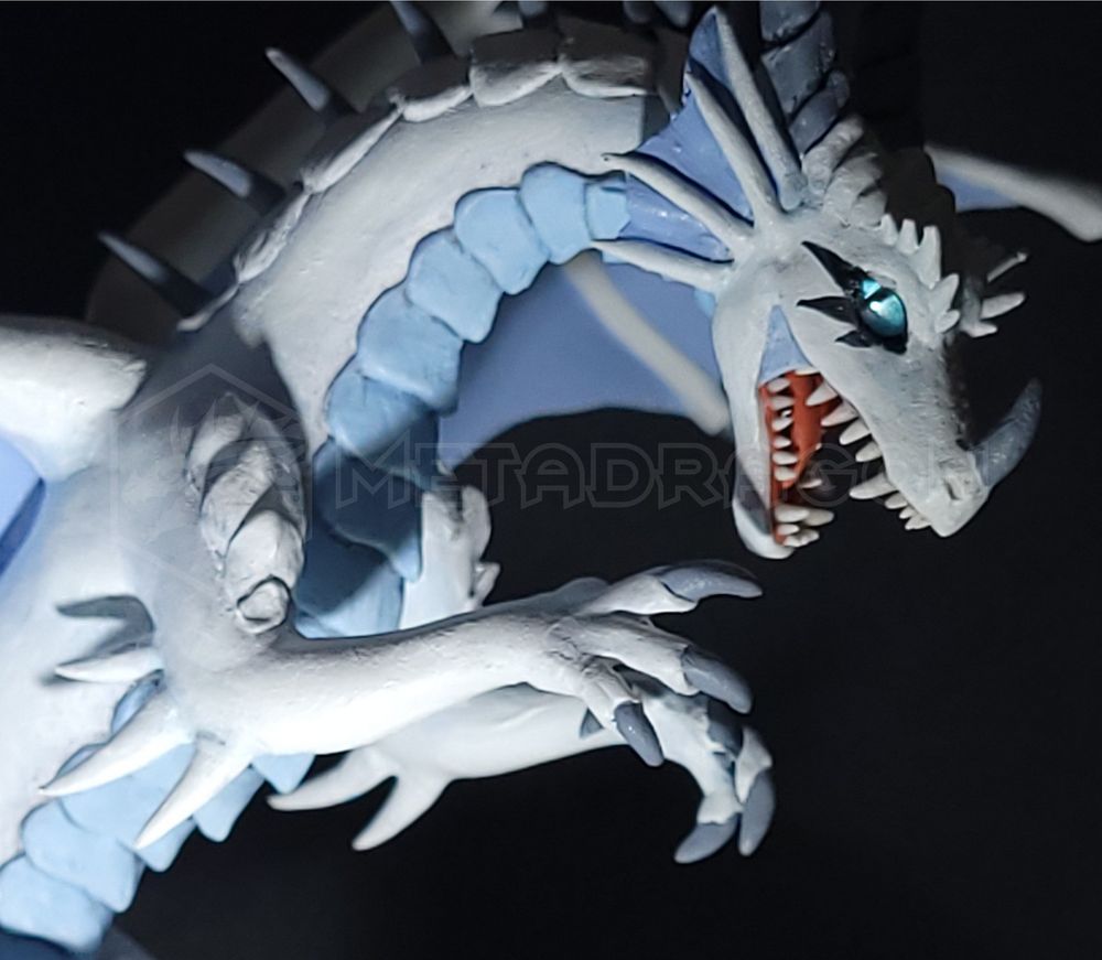 A modeled figurine of a white dragon sitting on a mountain ledge. Closeup view revealing blue eyes and a cheeky grin.