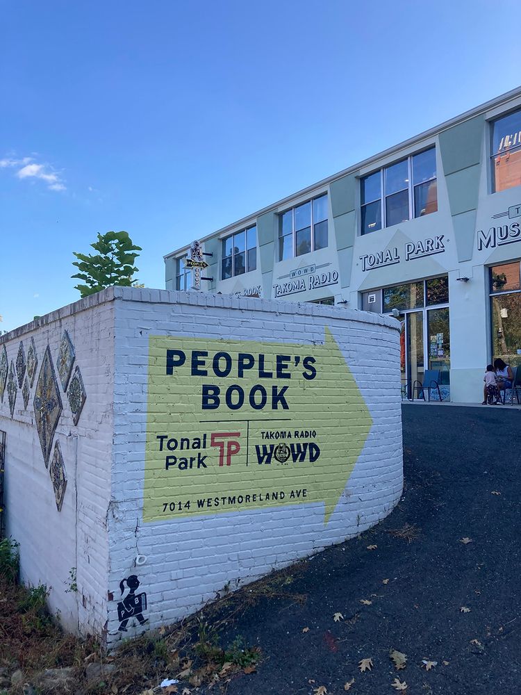 A large white painted brick wall leading into a parking lot under a blue evening sky. On the wall is a big painted yellow arrow with the signs People's Book, Tonal Park and Takoma Radio, 7014 Westmoreland Ave 