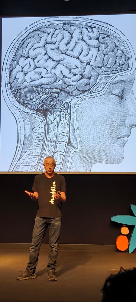 Una imagen de la charla: en la pantalla un dibujo de un cerebro dentro de una persona que está pintada de perfil. Delante y abajo el conferenciante vestido con camiseta negra
