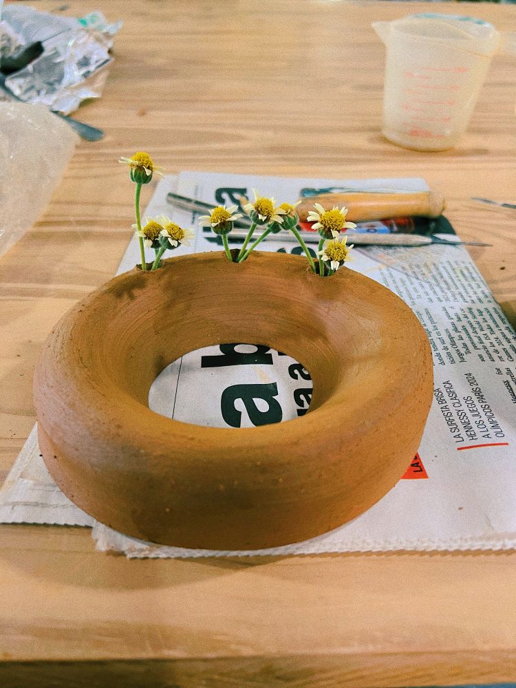 Fotografía de perfil que muestra una dona de cerámica, es un jarrón en forma de dona. La pieza está cruda (necesita la primera cocción al horno) y tiene tres huecos de los que salen unas pequeñas flores que parecen margaritas 