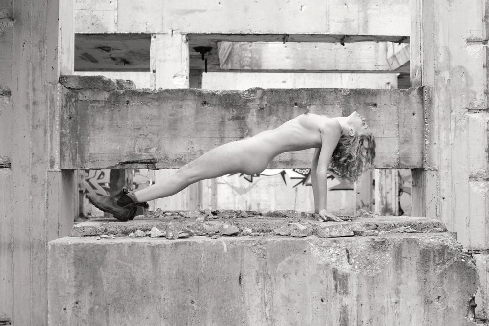 A nude model poses in boots doing a reverse press up on a concrete block in a ruinnned building