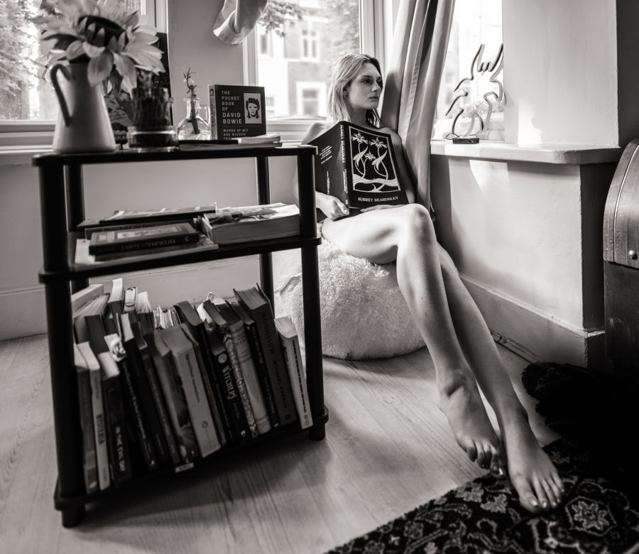 A model in her sitting room with a book case holds a book over her chest - her legs point towards the camera a wide angle lens distorts them making them appear impossibly long