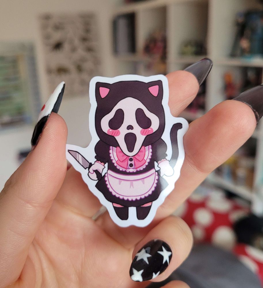 A hand holding up a small sticker of Ghostface is a maid dress with cat ears and a tail.