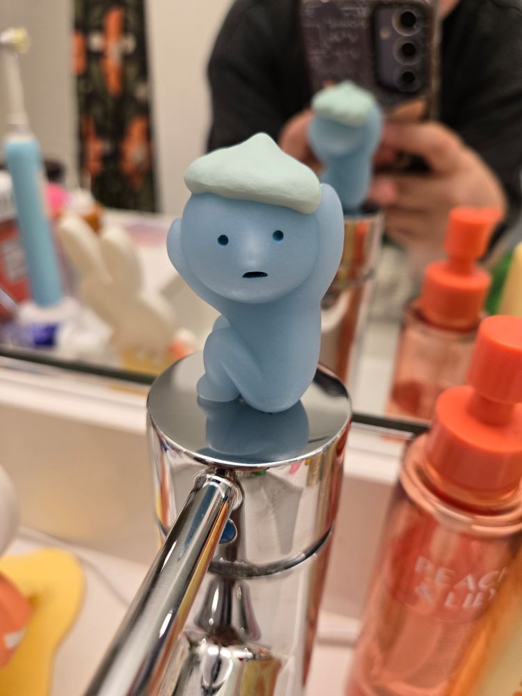 A photo blue smiski figurine washing his hair, he has a hat made of bubbles/lather. He is glued onto a sink faucet handle.