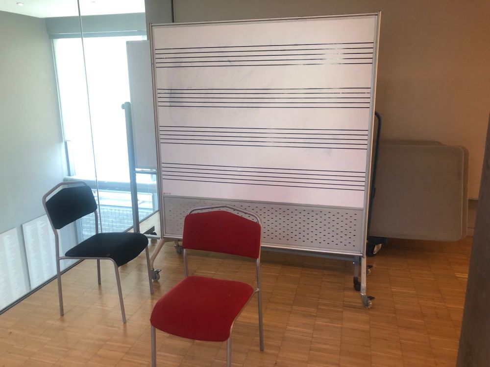 A large whiteboard with four bars of musical staff. A red chair and a black chair are in front of the whiteboard.
