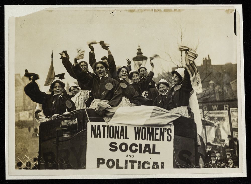 A monochrome photograph of a group of suffragettes dressed in mock prison garb, standing and waving inside a bus. The bus has a poster from the Women's Social and Political Union displayed on the front.