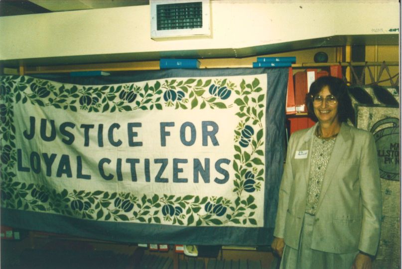 Rita Pankhurst smiling and stood next to a banner with "Justice For Loyal Citizens" on it.