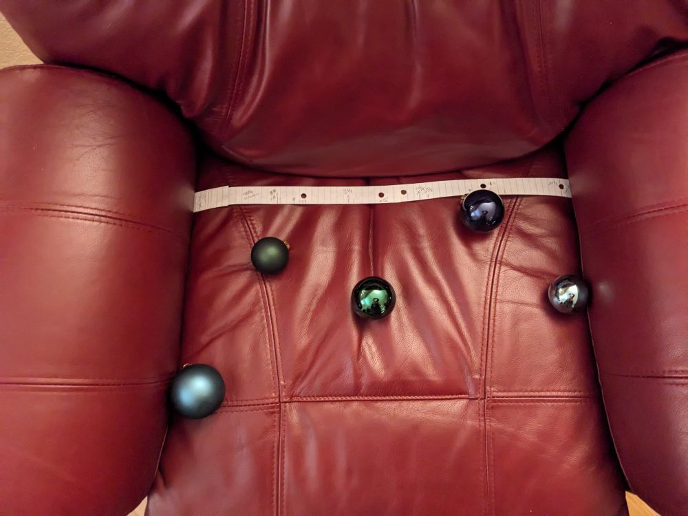 Photo of a maroon armchair with five baubles in different shades of green and blue on the seat, spaced out neatly with a piece of paper above them with markings to show proper placement.