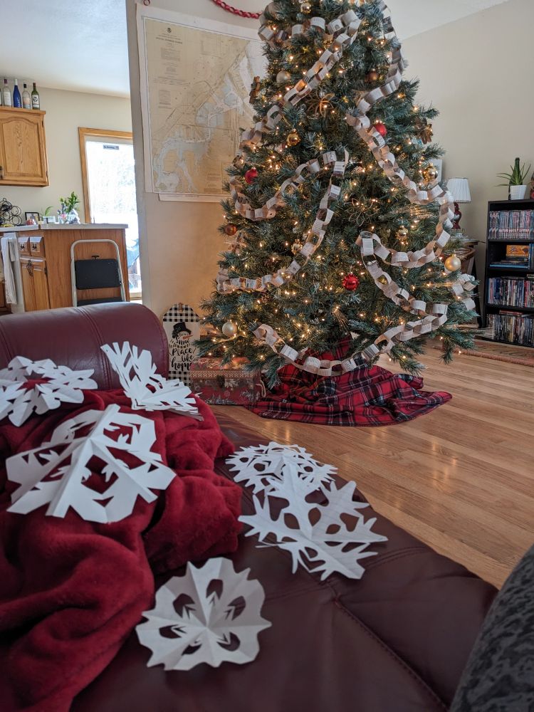 A photo of a Christmas tree inside a living room, decorated with newspaper chain garland, white lights, and red, gold, and white baubles. In the foreground on a maroon couch lie several freshly cut white paper snowflakes.