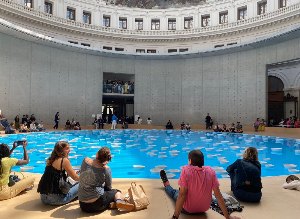 Plusieurs personnes au bord d‘un bassin bleu piscine sur lequel flottent des bols blancs 
