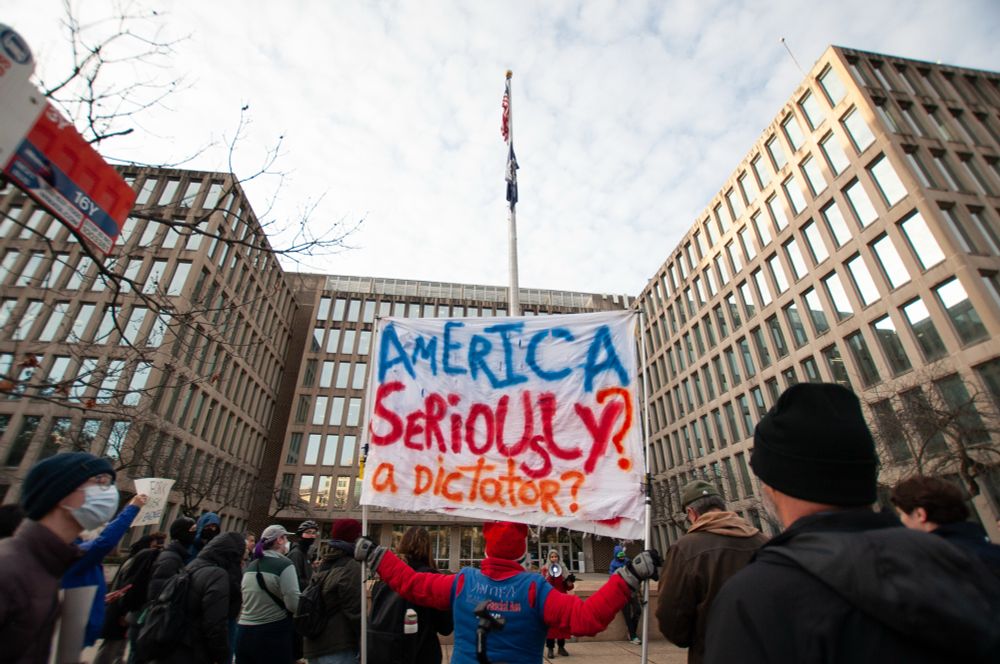 Wide photo of a crowd gathering in front of the federal building and the person in the center is holding a large banner that reads “America seriously? A dictator?”