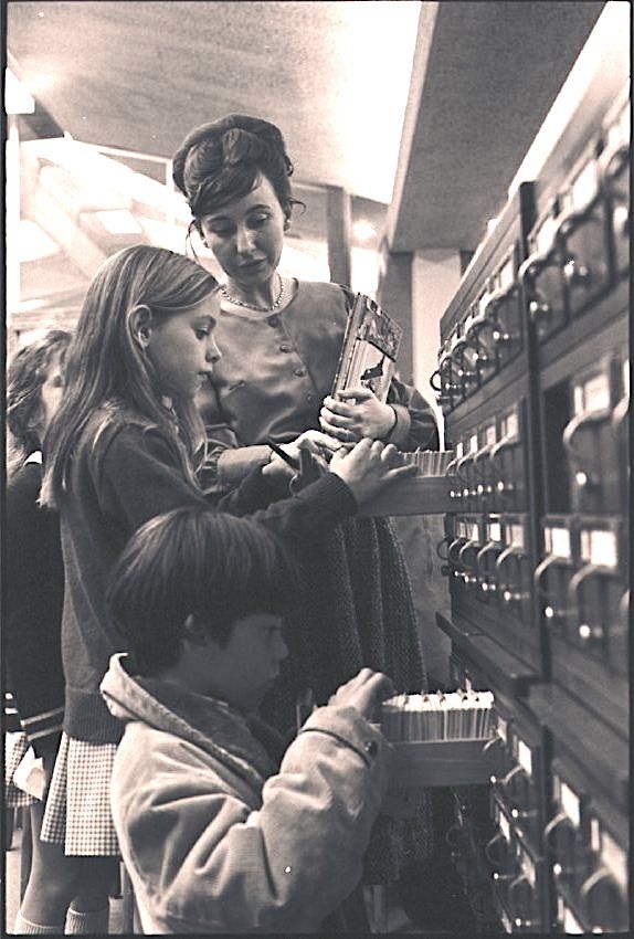 Children searching through library card catalog as librarian looks on.