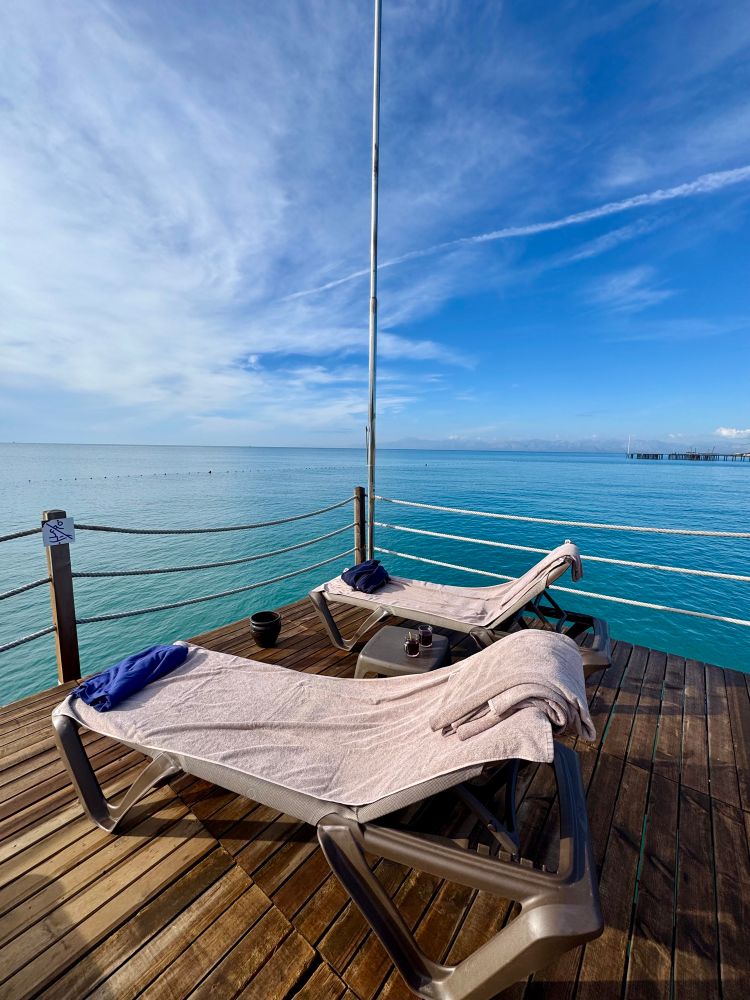 Zwei Liegestühle mit Handtüchern stehen auf einem hölzernen Steg direkt über dem türkisblauen Meer. Zwischen ihnen steht ein kleiner Tisch mit zwei Getränken, während der Himmel darüber weit und wolkig wirkt. Die Szene vermittelt Ruhe und Urlaubsstimmung mit offenem Blick auf das Wasser und den Horizont.
