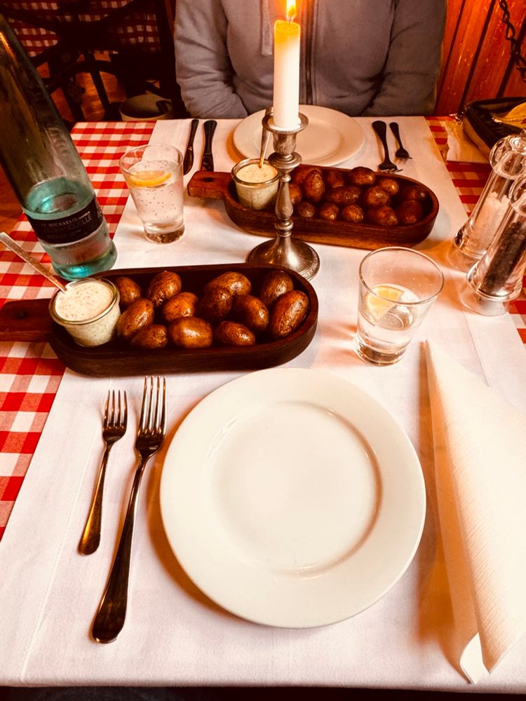 Ein gemütlich gedeckter Tisch mit rot-weißem Tischtuch.
In der Mitte brennt eine Kerze im silbernen Kerzenhalter.
Zwei Holzschalen mit kleinen Kartoffeln und Dip stehen bereit.
Eine Flasche Mineralwasser und Gläser mit Zitronenscheibe sind zu sehen.
Besteck und Teller liegen ordentlich vor jedem Platz.
Eine Person mit grauer Jacke sitzt im Hintergrund.