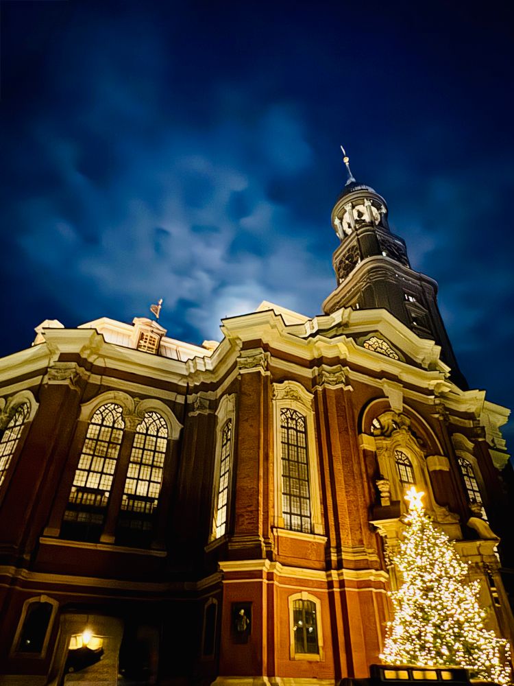Das Bild zeigt eine prachtvoll beleuchtete Kirche bei Nacht, deren warme Lichtakzente stark gegen den tiefblauen Himmel kontrastieren. Die Architektur wirkt barock und imposant, mit hohen Fenstern und reich verzierten Fassadenelementen. Rechts im Vordergrund steht ein festlich geschmückter Weihnachtsbaum, der die Szene noch stimmungsvoller macht. Die dramatischen Wolken hinter dem Turm verstärken den Eindruck einer besonderen, fast feierlichen Atmosphäre. 