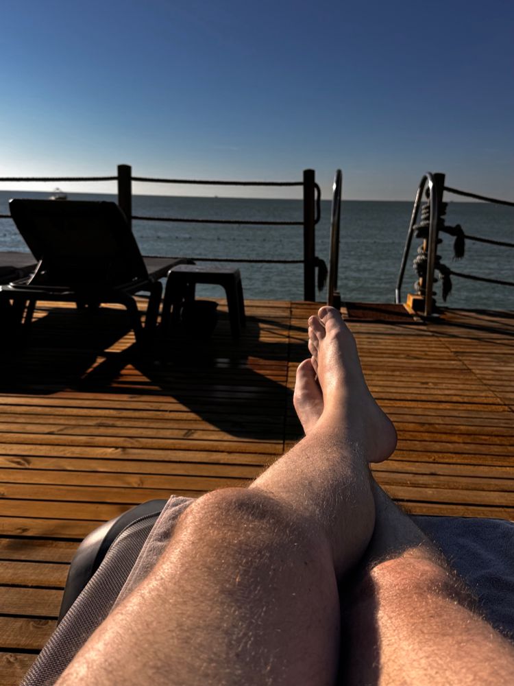 Eine Person liegt entspannt auf einer Liege und hat die Beine übereinandergeschlagen. Der Blick geht über einen hölzernen Steg hinaus aufs ruhige Meer. Im Hintergrund stehen weitere Liegen und eine Reling begrenzt den Steg. Die Sonne scheint, der Himmel ist klar und es herrscht eine ruhige, sommerliche Stimmung.