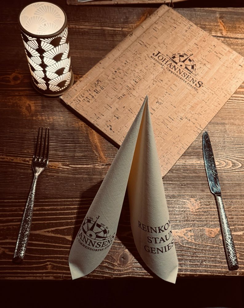 Der Holztisch ist stimmungsvoll mit einer filigranen Lampe, Besteck und einer kunstvoll gefalteten Serviette gedeckt.
Auf der Serviette steht der Name des Restaurants „Johannsen’s“ sowie ein einladender Schriftzug.
Rechts liegt eine Speisekarte mit Korkoptik und dem Logo des Hauses.
Das warme Licht der Lampe erzeugt eine gemütliche und elegante Atmosphäre.
Alles wirkt sorgfältig arrangiert und bereit für ein entspanntes Abendessen.