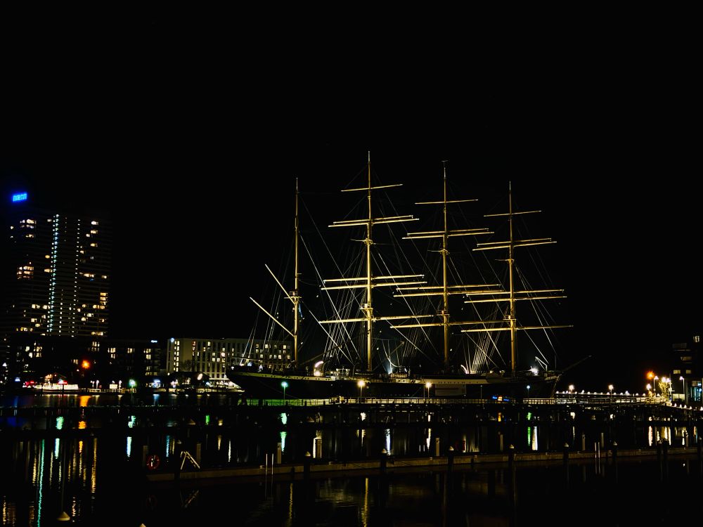 Ein beleuchtetes Segelschiff liegt nachts ruhig im Hafen, klar umrissen vor dem dunklen Himmel. Die Masten ragen symmetrisch empor und werden von starken Scheinwerfern angestrahlt. Auf dem Wasser spiegeln sich die Lichter der Umgebung in langen, farbigen Streifen. Im Hintergrund stehen moderne Gebäude, deren Fenster punktuell leuchten. Die Szene wirkt geordnet und technisch klar beleuchtet. Alles erscheint ruhig, ohne besondere Bewegung. Die Kombination aus Schiff und Stadt vermittelt einen sachlichen, nüchternen Eindruck.