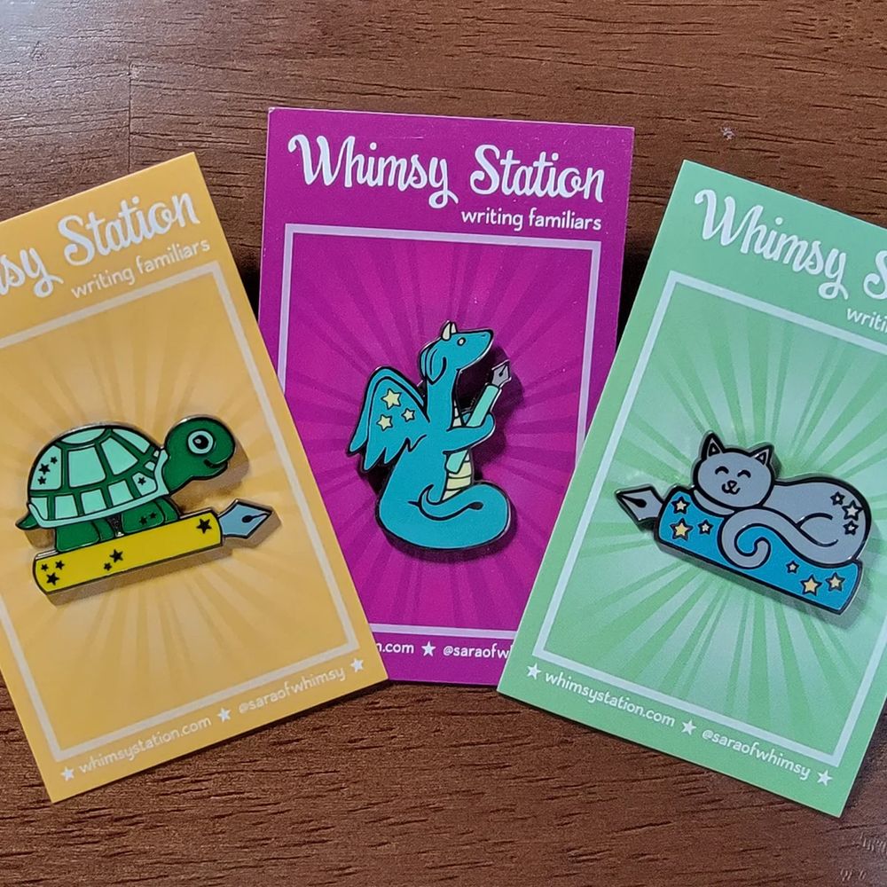 A trio of writing familiar enamel pins on their backing cards.  A turtle, a dragon, and a cat each with a fountain pen.