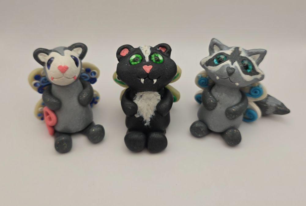Three fairy creatures: an opossum, a skunk, and a raccoon, each with heart shaped noses and fangs, facing the camera.  