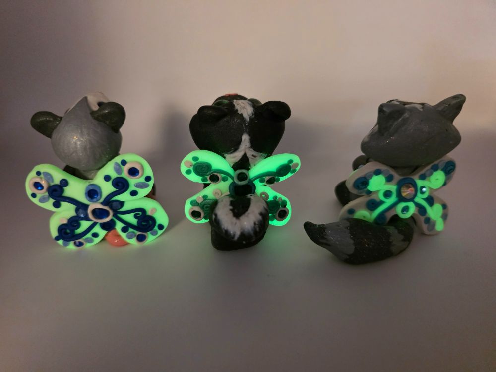 Three fairy creatures: an opossum, a skunk, and a raccoon, from behind showing their wings with rhinestones and swirling designs.  Showing their glowing wings.