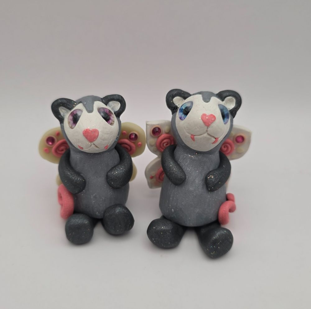 Two little fairy opossums facing forward with swirly tails, heart shaped noses, wings, colorful eyes, and pink fangs.