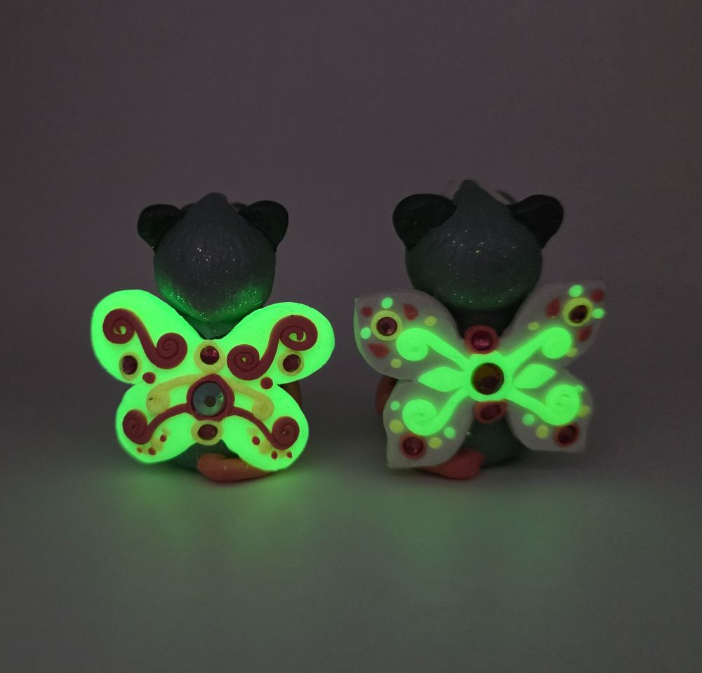 Two little fairy opossums from behind, showing off their swirly wings decorated with rhinestones and swirls, in the dark showing them glowing